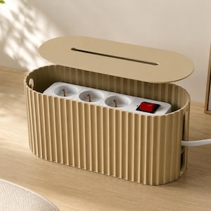 May include: A beige, oval-shaped cable management box with a ribbed exterior texture and a removable lid. Inside, a white power strip with three electrical outlets and a red illuminated power switch is visible, with a white cord extending from the side.