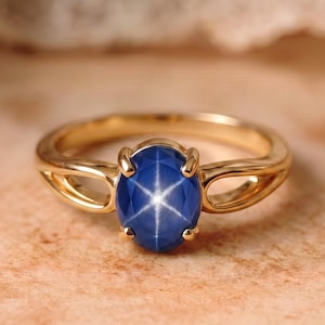 May include: A gold ring featuring a central oval-shaped blue star sapphire gemstone. The stone displays a distinct six-rayed star pattern on its surface. The band is crafted from polished yellow gold and has a split shank design holding the stone.