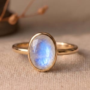 Puede incluir: Un anillo de oro con una piedra lunar central de forma ovalada y superficie facetada. La piedra lunar muestra un aspecto translúcido con una distintiva adularescencia azul. La piedra está engastada en un bisel de oro liso.