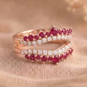 May include: A rose gold ring set featuring two curved bands. The top band displays a row of round red rubies and a row of clear white diamonds. The bottom band mirrors this design with a row of clear white diamonds and a row of round red rubies.