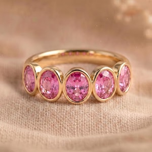 Peut inclure: Une bague en or ornée de cinq pierres précieuses roses de taille ovale disposées en rangée. Les pierres sont alignées horizontalement sur le devant de l'anneau, chacune fixée dans un serti clos en or lisse qui souligne la couleur rose vive.