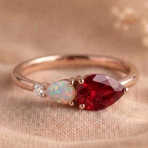 Könnte beinhalten: Ein Ring aus Roségold mit einem birnenförmigen roten Edelstein, einem birnenförmigen schillernden Opal und einem kleinen runden klaren Stein. Die Steine sind in einer Reihe auf der Ringschiene angeordnet und bilden ein Cluster-Design.