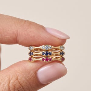 Può includere: Una pila di tre anelli in oro con design a fascia ondulata. L'anello superiore presenta pietre chiare, quello centrale pietre blu scuro e quello inferiore pietre rosa brillante, il tutto incastonato in una delicata montatura metallica.