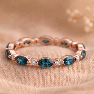 May include: A rose gold eternity band featuring alternating marquise-cut blue gemstones and small round white stones. The ring has a continuous circular design with a polished metal finish, showcasing a repeating pattern of blue and white stones.