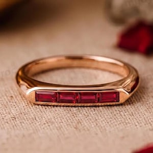 Op de afbeelding: Een roségouden ring met vier rechthoekige, in baguette geslepen rode robijnen die horizontaal op een rij in het midden van de band zijn gezet. Het metaal heeft een gepolijste afwerking en de stenen zijn levendig rood van kleur.