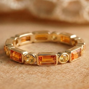 Puede incluir: Un anillo de oro con un patrón alterno de piedras preciosas naranjas de corte rectangular baguette y piedras preciosas amarillas de corte redondo brillante, engastadas en una fila continua alrededor de toda la banda.