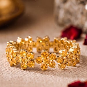 Könnte beinhalten: Ein goldfarbener Ring mit einem durchgehenden Band aus miteinander verbundenen blumenförmigen Motiven. Jede Blume besteht aus kleinen, runden, gelb-orangen Edelsteinen in einer Metallfassung, die ein sich wiederholendes Blumenmuster bilden.