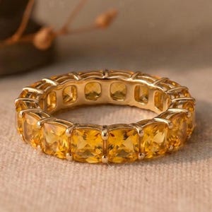 Peut inclure: Une alliance en or ornée d'une rangée continue de pierres précieuses citrines jaunes de taille carrée, fixées par des griffes. La bague présente un agencement uniforme de pierres facettées qui entourent tout l'anneau, révélant une teinte chaude.