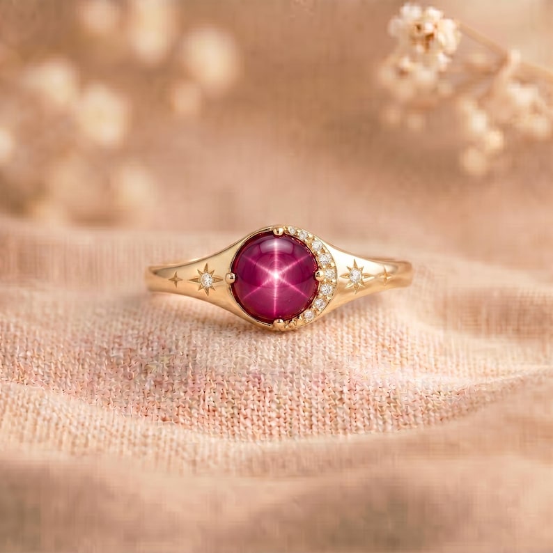 May include: A gold ring featuring a round, deep pink star sapphire cabochon at the center. The band is accented with a crescent moon shape set with small white diamonds and engraved star motifs on either side of the central gemstone setting.