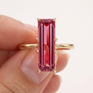 May include: A gold ring featuring a long, rectangular, baguette-cut pink gemstone set vertically. The stone is held in place by eight small gold prongs, four on each side, and the band is a simple, polished gold metal finish.