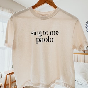 May include: A cream-colored short-sleeve t-shirt featuring the black text "sing to me paolo" printed in a serif font on the center of the chest. The shirt is displayed on a wooden hanger against a neutral background.