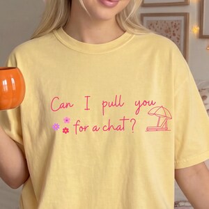 May include: A pale yellow short-sleeve t-shirt featuring the pink text Can I pull you for a chat? accompanied by small pink and purple flower icons and a pink line-art illustration of a beach lounge chair with an umbrella.
