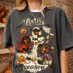 Puede incluir: Una camiseta gris carbón con un gráfico de tres perros salchicha con disfraces de Halloween, incluyendo un diablo, un fantasma y una bruja. El texto 'tis the season' aparece en cursiva, rodeado de calabazas, murciélagos y telarañas.