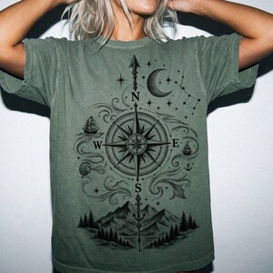 Peut inclure: Un t-shirt à manches courtes vert sauge avec une impression graphique noire représentant une rose des vents centrale avec les directions N, S, E et O. Le motif inclut un croissant de lune, des étoiles, un voilier, une ancre et des montagnes.