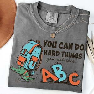 May include: A charcoal gray t-shirt featuring a graphic of a green alligator carrying a blue and orange backpack. The text reads, "YOU CAN DO HARD THINGS you got this!" along with the letters "ABC" in orange and blue. The shirt has a crew neck.