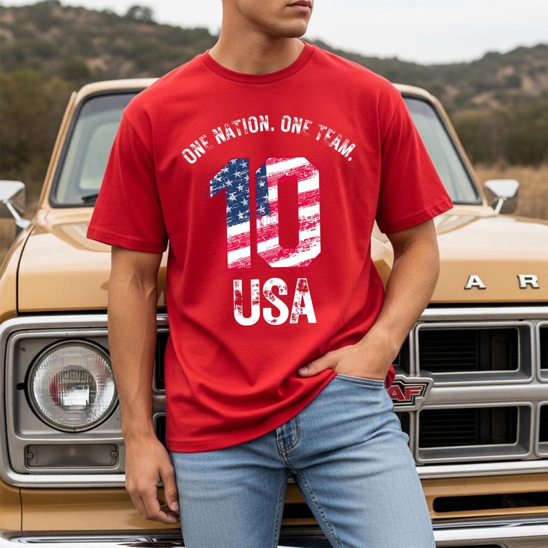May include: A red short-sleeved t-shirt featuring the text ONE NATION, ONE TEAM, arched above a large number 10 filled with a distressed American flag pattern, with the word USA printed in white below the number in a matching distressed font.