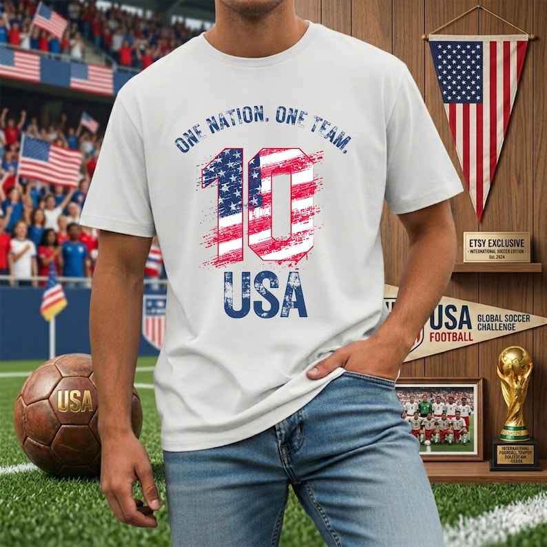 May include: A white short-sleeved t-shirt featuring the text ONE NATION. ONE TEAM. above a large number 10 filled with a red, white, and blue American flag pattern, with the word USA printed in blue below the number.