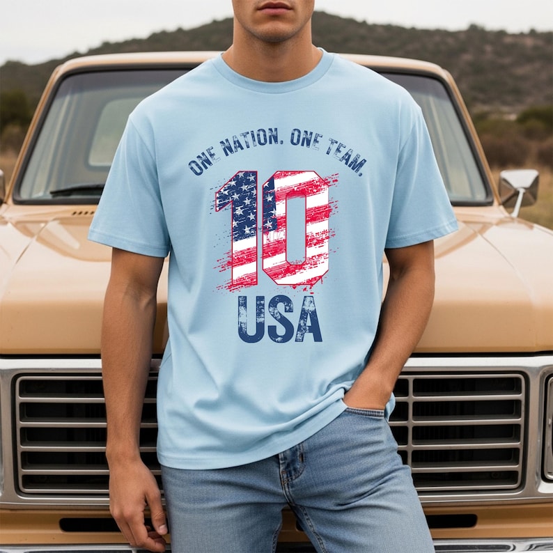 May include: A light blue short-sleeve t-shirt featuring the text ONE NATION. ONE TEAM. above a large number 10 filled with a red, white, and blue American flag pattern, with the word USA printed in blue below the number in a distressed font.