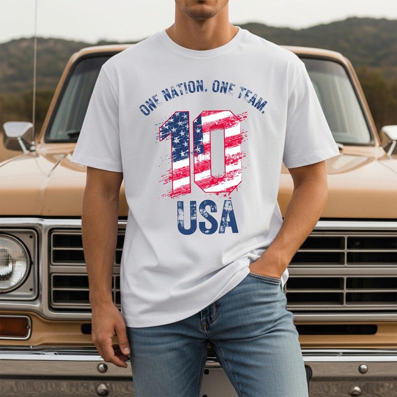May include: A white short-sleeved t-shirt featuring the text ONE NATION. ONE TEAM. in blue, with a large number 10 filled with a red, white, and blue American flag pattern, and the word USA in blue text positioned below the number 10.