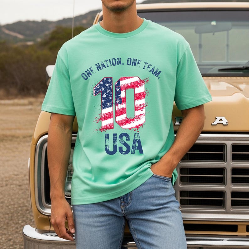 May include: A mint green short-sleeve t-shirt featuring the text ONE NATION. ONE TEAM. in dark blue above a large number 10 filled with a red, white, and blue American flag pattern. The word USA is printed in dark blue below the number 10.