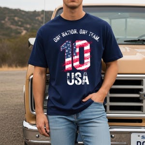May include: A navy blue short-sleeved t-shirt featuring a graphic print with the text ONE NATION, ONE TEAM, and USA. The number 10 is displayed in the center, filled with a red, white, and blue American flag pattern with a distressed texture.