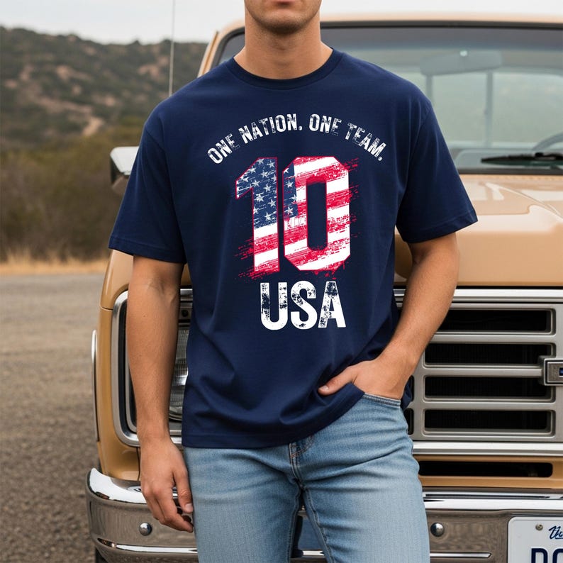 May include: A navy blue short-sleeved t-shirt featuring a graphic print with the text ONE NATION, ONE TEAM, and USA. The number 10 is displayed in the center, filled with a red, white, and blue American flag pattern with a distressed texture.