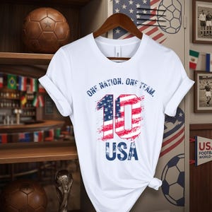 May include: A white short-sleeved t-shirt featuring the text 'ONE NATION, ONE TEAM,' the number '10' filled with a red, white, and blue American flag pattern, and the word 'USA' in blue, all displayed in a distressed, weathered graphic style.
