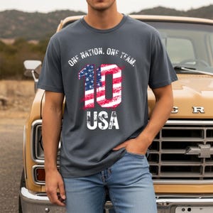 May include: A charcoal gray short-sleeve t-shirt featuring the white text ONE NATION. ONE TEAM. above a large number 10 filled with a red, white, and blue American flag pattern, with the word USA printed in white below the number 10.