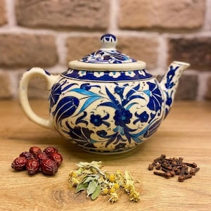 May include: A ceramic teapot with a blue and white floral design. The teapot has a curved handle and spout. Dried rosehips, herbs, and cloves are arranged near the teapot on a wooden surface. The background features a brick wall.