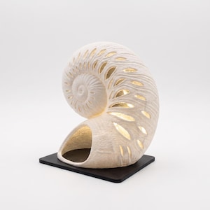 May include: A decorative, cream-colored seashell-shaped lamp with a spiral design and cut-out patterns. The interior is illuminated with a warm, golden light. It sits on a small, black square base, creating a unique home decor accent.