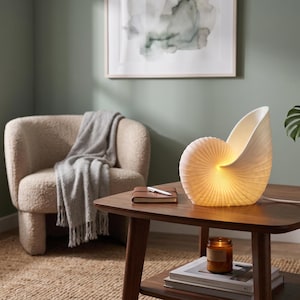 May include: A white, nautilus shell-shaped table lamp with a ribbed texture and a warm, internal glow. The lamp sits on a wooden table, connected by a white power cord, emitting soft light from its curved, spiral structure.