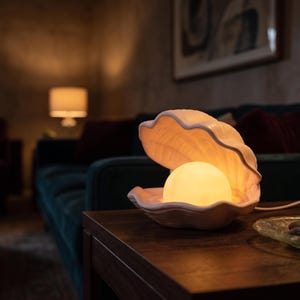 Op de afbeelding: Een keramische tafellamp in de vorm van een open schelp met een zacht gloeiende, bolvormige lamp erin. De schelp heeft een bleke, neutrale kleur met een getextureerd, geribbeld oppervlak en is verbonden met een wit netsnoer.