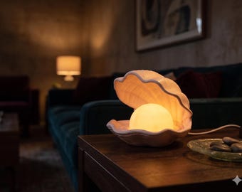 Könnte beinhalten: Eine Keramik-Tischleuchte in Form einer geöffneten Muschel mit einer sanft leuchtenden, kugelförmigen Glühbirne im Inneren. Die Muschel hat eine helle, neutrale Farbe mit einer texturierten, gerippten Oberfläche und ein weißes Stromkabel.