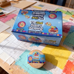 May include: A blue box and a round bamboo container with a label. The box reads "The Viral Mystery Dumpling!" with colorful dumpling illustrations. The container has a label that says "The Viral Mystery Dumpling!" and "What Color Will You Get?"