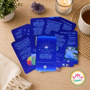 May include: A deck of blue "Wellness Cards" for cleaning and organizing, with various tips on each card. The cards are arranged on a wooden surface, alongside a small plant, a candle, and a cup of tea.