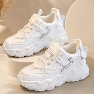 May include: Pair of white chunky sneakers with a thick sole. The sneakers have a lace-up closure and a strap detail on the back. The design features a wave-like pattern on the side.