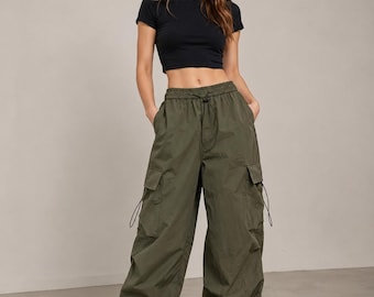 May include: A woman wearing a black crop top and olive green cargo pants with multiple pockets and drawstring details. The pants are loose-fitting and paired with beige sneakers. The outfit is casual and stylish.