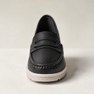 May include: Black leather loafer with a textured upper, a decorative strap, and a light-colored sole. The shoe has a classic design with a modern twist, suitable for various occasions.