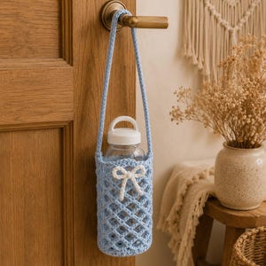May include: A light blue crocheted water bottle holder with a diamond-shaped mesh pattern and a small white crocheted bow detail. The holder features a long matching strap and is hanging from a wooden door handle, containing a clear plastic bottle.