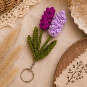 May include: A crochet keychain featuring two lavender-like flower stalks in deep purple and light lilac, attached to a green stem with several pointed leaves. The floral element is connected to a circular silver-toned metal split ring.