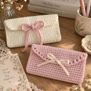May include: Two rectangular crochet pouches, one cream-coloured with a pink yarn bow and one light pink with a cream-coloured ribbon bow. Both pouches feature a flap closure and a textured stitch pattern, presented as small envelope-style cases.