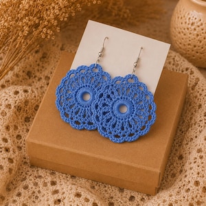 May include: A pair of round, bright blue crochet earrings featuring a detailed circular lace pattern with a central open hole. The earrings are attached to silver-toned metal fishhook wires and are displayed on a white rectangular card.