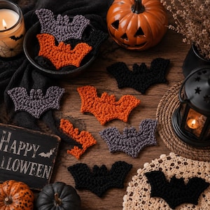 May include: A collection of crocheted bat appliques in black, orange, and dark gray yarn. The bats are scattered across a wooden surface alongside a ceramic pumpkin, a black lantern, a candle in a glass jar, and a sign that reads Happy Halloween.