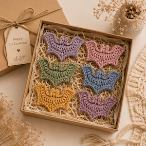 May include: A set of six crocheted bat ornaments in purple, pink, olive green, blue, mustard yellow, and lavender, arranged inside a square cardboard gift box with a tag that reads happy halloween and a small heart icon, resting on a light surface.