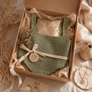 Op de afbeelding: Een saliegroene gehaakte top met brede bandjes, gepresenteerd in een kartonnen geschenkdoos bekleed met vloeipapier. Het kledingstuk is versierd met een beige lint, een klein rond gehaakt accent en een takje gedroogde bloemen aan de voorzijde.