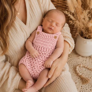 May include: A light pink crocheted baby romper featuring a textured stitch pattern, a square neckline, and two small wooden buttons at the shoulder straps. The garment is sleeveless and has a fitted bodice with a slightly flared lower section.