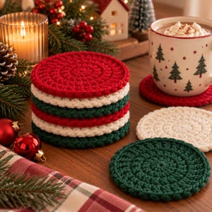 May include: A stack of round, crocheted coasters in alternating red, white, and dark green colours. Two additional individual coasters, one white and one dark green, lie flat on the surface next to a ceramic mug decorated with green pine tree patterns.