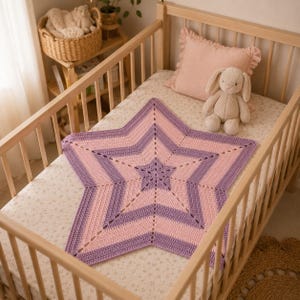 Könnte beinhalten: Eine sternförmige Häkeldecke mit abwechselnden Streifen aus hellrosa und lavendelfarbenem Garn. Die Decke liegt flach auf einer Kinderbettmatratze, begleitet von einem Plüschhasen und einem hellrosa Kissen mit Rüschen in einem Holzbett.