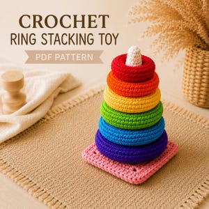 May include: A crochet ring stacking toy PDF pattern featuring a stack of six colorful rings in red, orange, yellow, green, blue, and purple, topped with a white cone, all resting on a square pink base with a textured, handmade yarn finish.