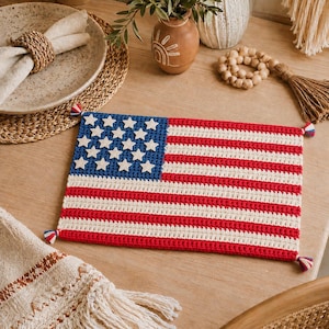 May include: A rectangular crochet placemat featuring the design of the American flag with horizontal red and white stripes and a blue canton containing small white stars. The corners of the placemat are finished with small red, white, and blue tassels.